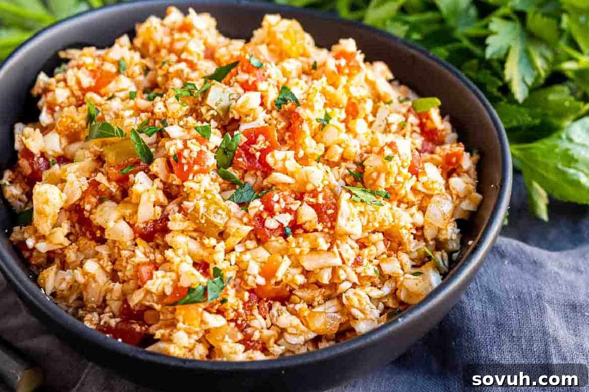 A black bowl filled with a dish of cauliflower rice mixed with diced tomatoes, herbs, and spices. Garnished with fresh parsley, resting on a fabric surface with greenery in the background.