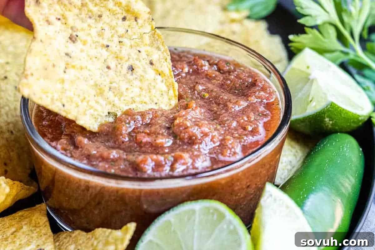 A tortilla chip is dipped into a bowl of red salsa surrounded by lime halves, a jalapeño, and cilantro.