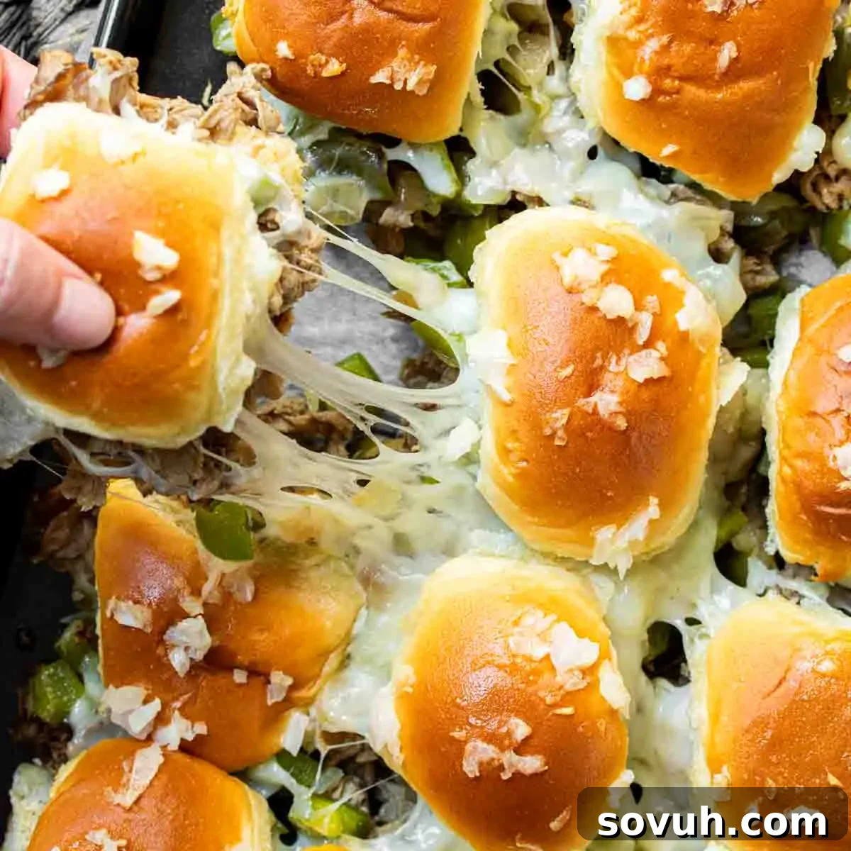 A person removing freshly baked Philly Cheesesteak Sliders from a pan, highlighting their golden-brown tops and melted cheese filling.