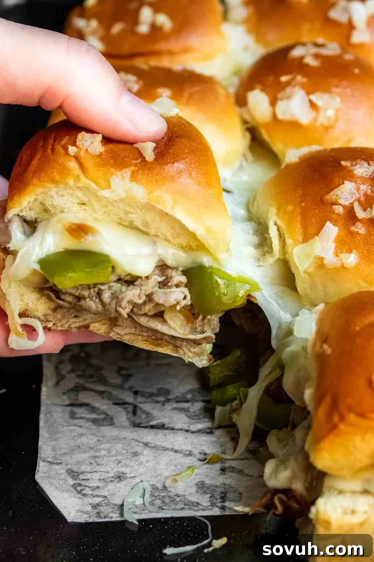 A hand reaching for a Philly Cheesesteak Slider from a baking dish, showcasing the melted cheese.