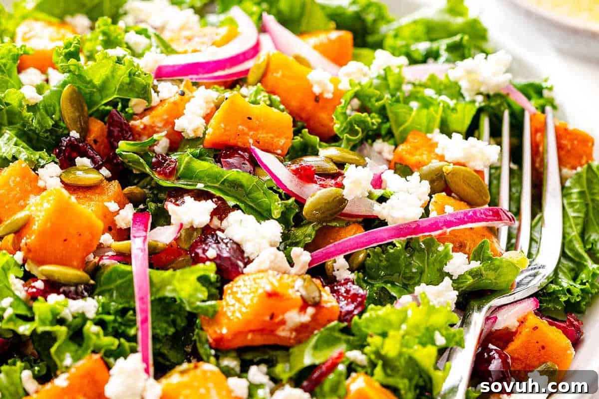 A delightful close-up of a hearty Sweet Potato Salad, showcasing vibrant curly kale, tender roasted sweet potato cubes, thinly sliced red onion, crumbly feta cheese, plump cranberries, and crunchy pumpkin seeds (pepitas). A fork rests invitingly on the side, inviting a taste.