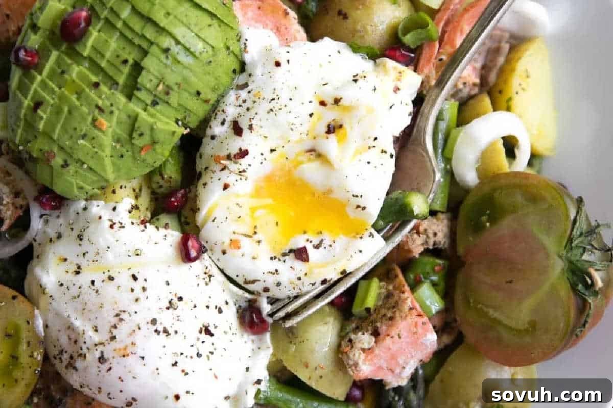 A gourmet salad featuring perfectly poached eggs, creamy avocado slices, vibrant cherry tomatoes, crunchy croutons, flakes of tender salmon, ruby-red pomegranate seeds, and fresh green onions, elegantly seasoned with cracked pepper.