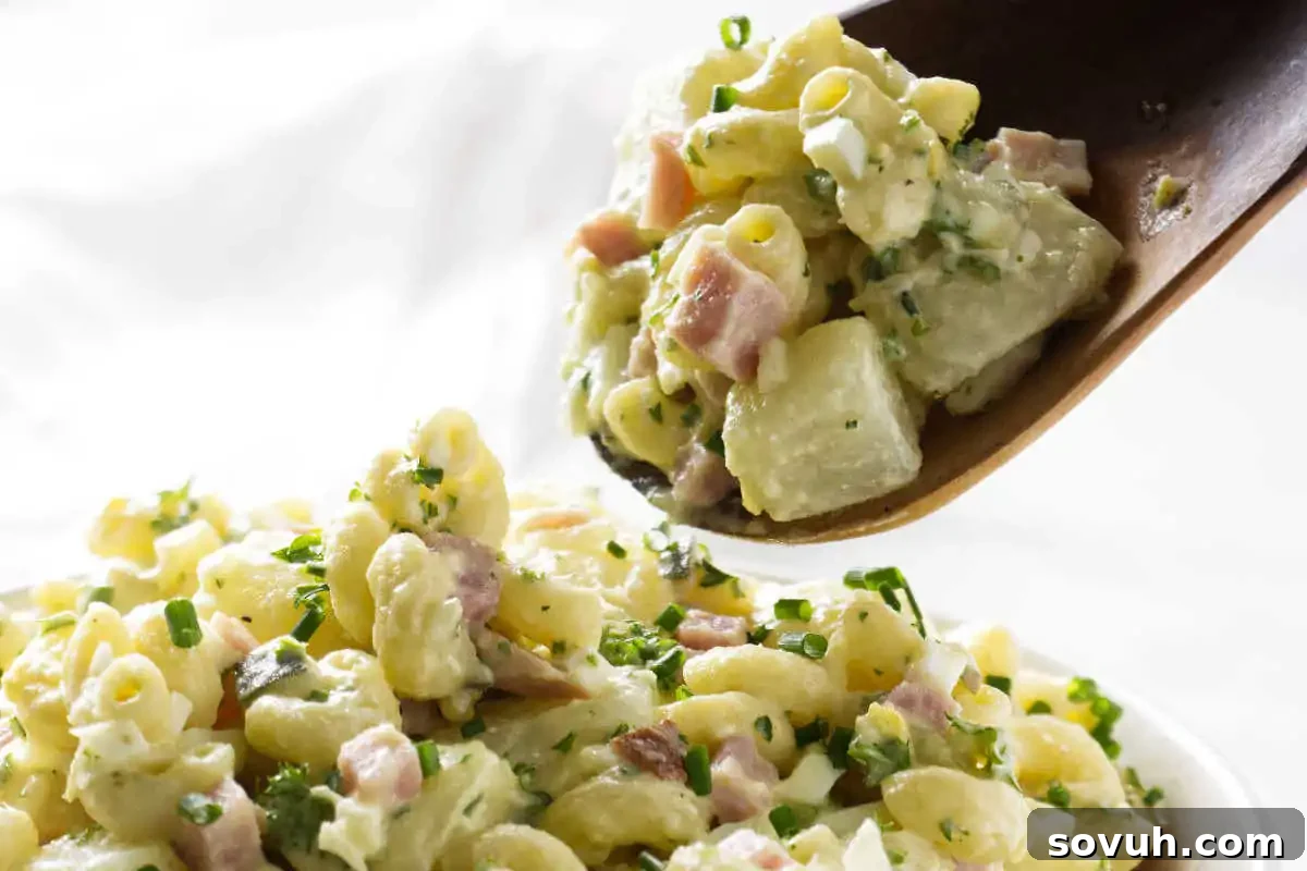 A wooden spoon scooping out a generous portion of creamy Potato Mac Salad, richly mixed with ham, crisp celery, and fresh chives from a large serving bowl.