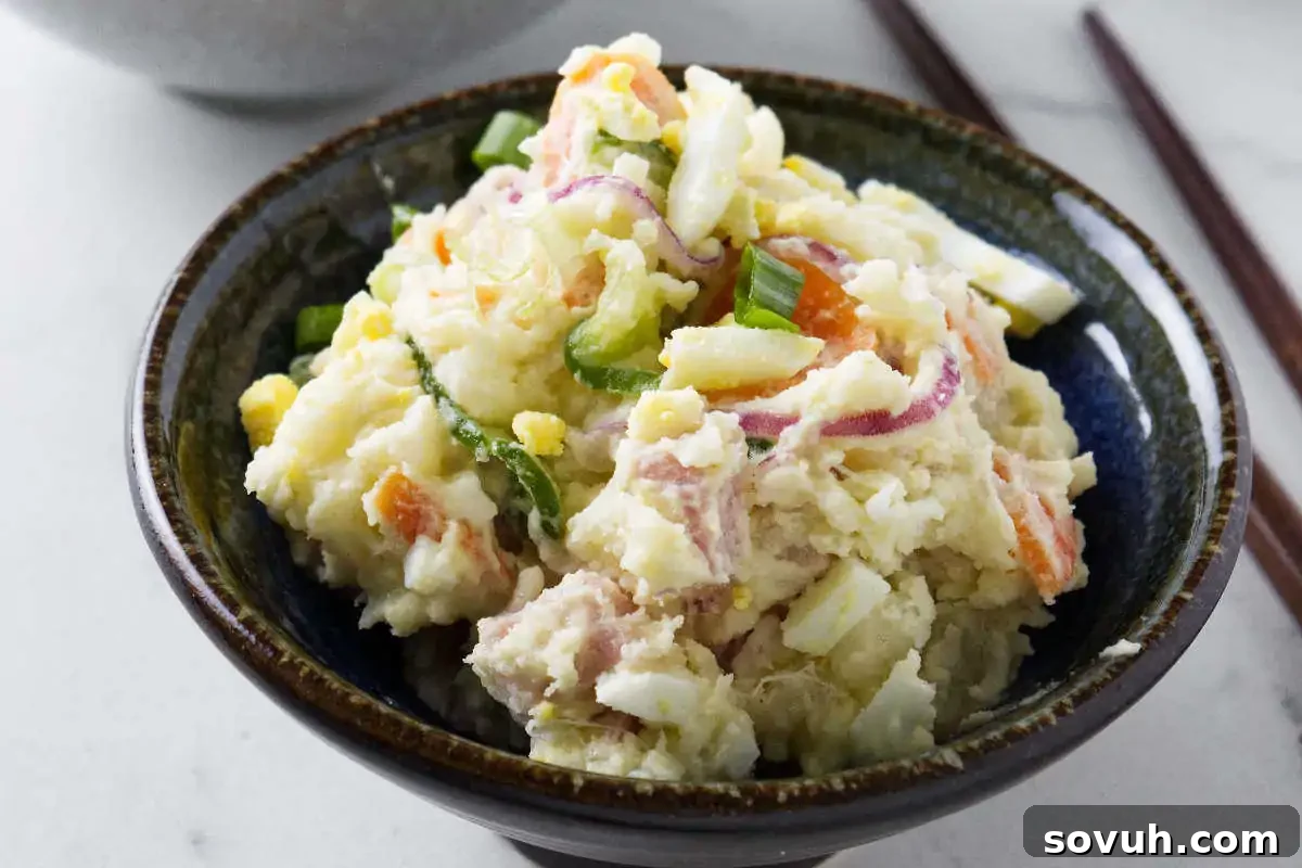 A beautifully presented bowl of Japanese Potato Salad, featuring creamy mashed potatoes, delicate slices of onion and carrot, and vibrant green onions, served in a dark ceramic bowl on a light background, with chopsticks nearby.
