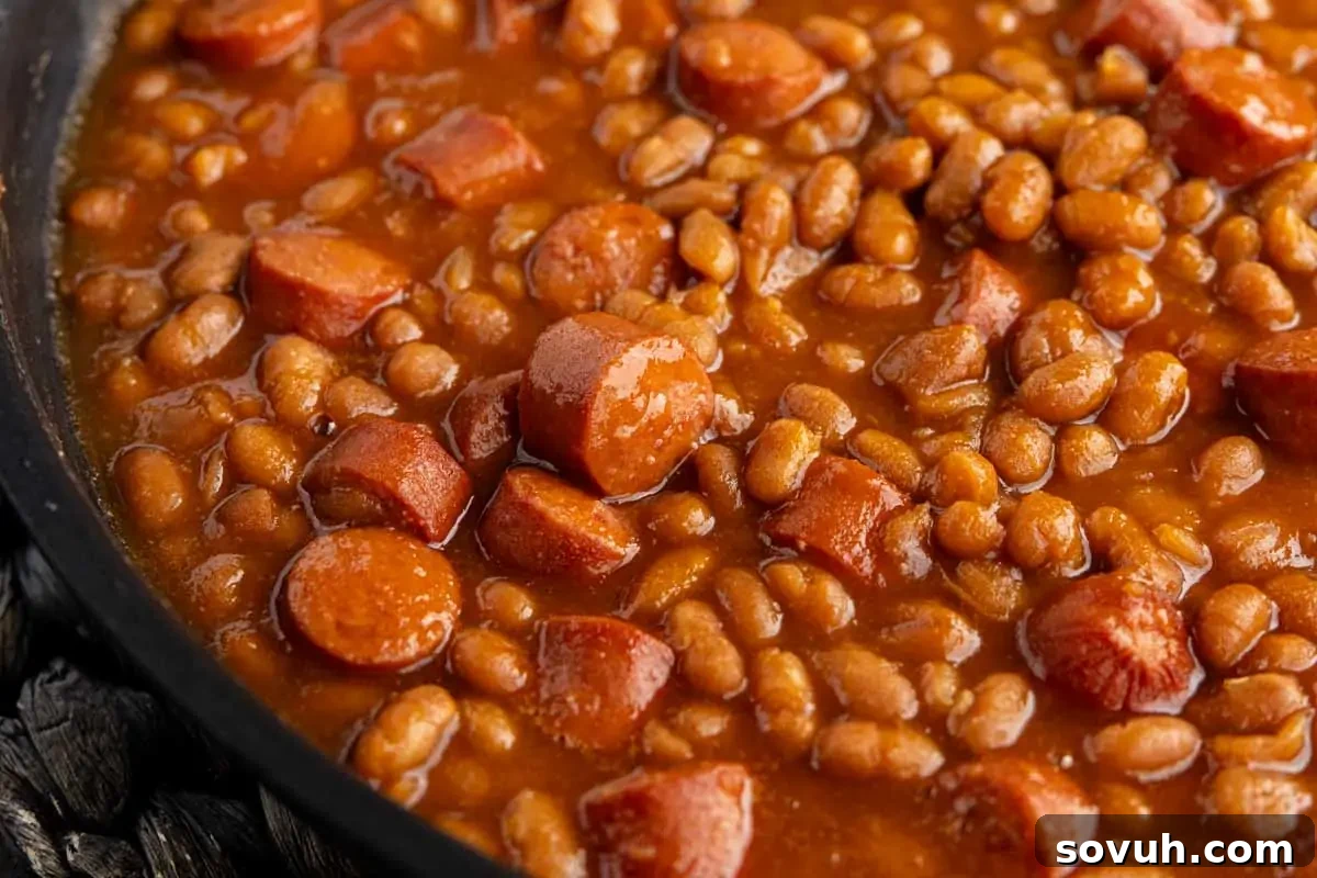 Close-up of baked beans mixed with sliced hot dogs in a brown sauce, shown in a pan.