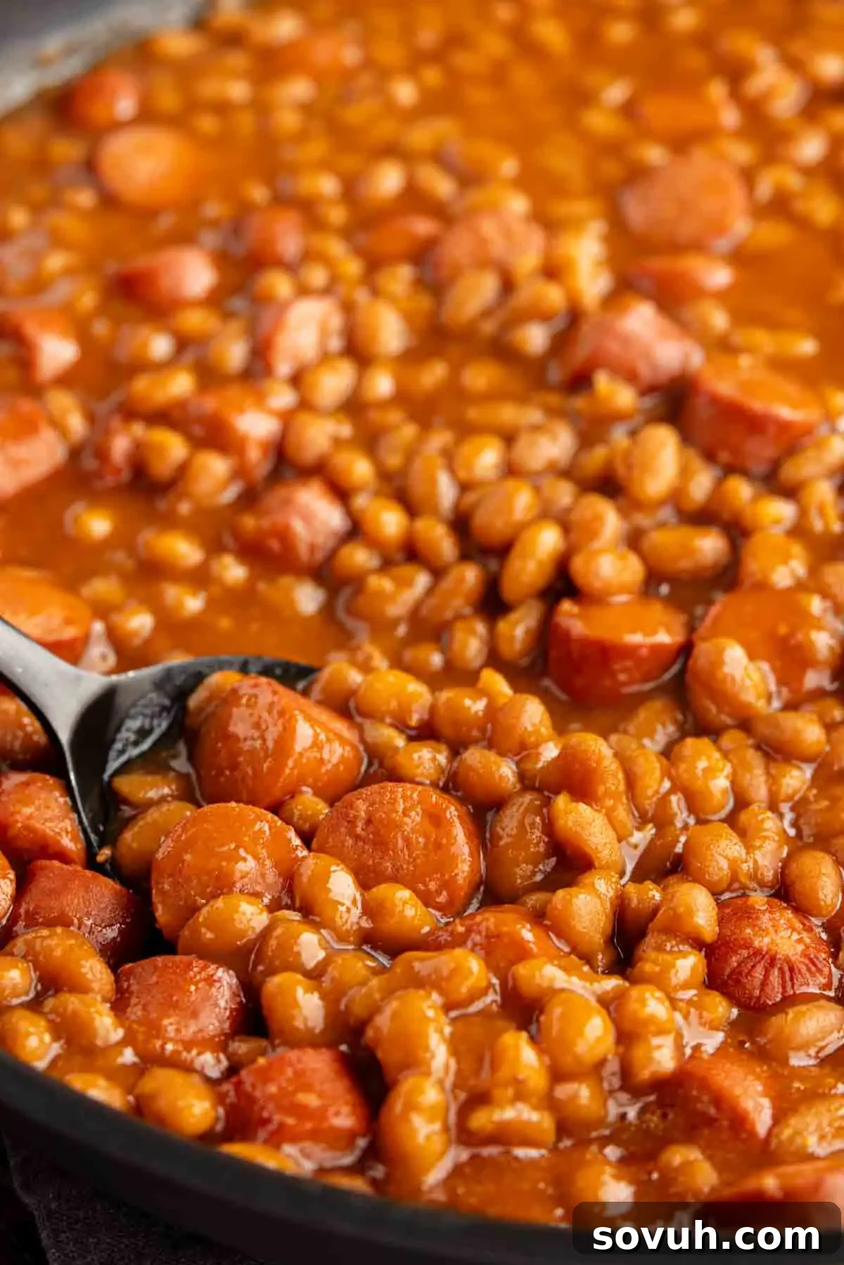 A skillet filled with franks and beans. A spoon rests in the mixture.
