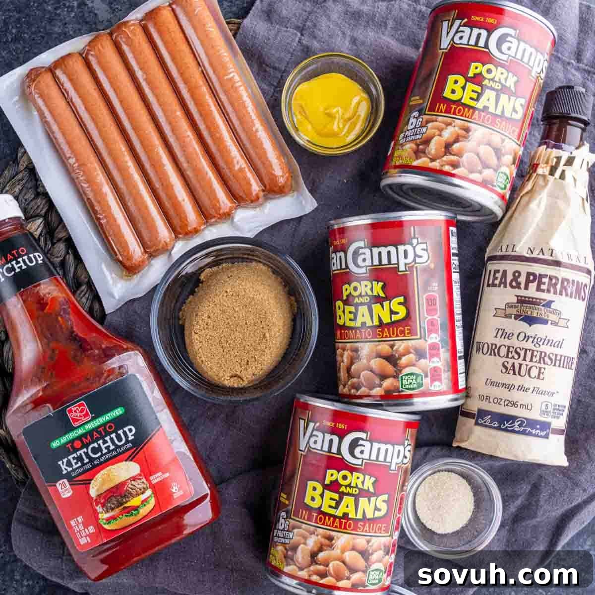 Ingredients for a dish include hot dogs, cans of pork and beans, ketchup, Worcestershire sauce, brown sugar, mustard, and seasoning set on a dark cloth.