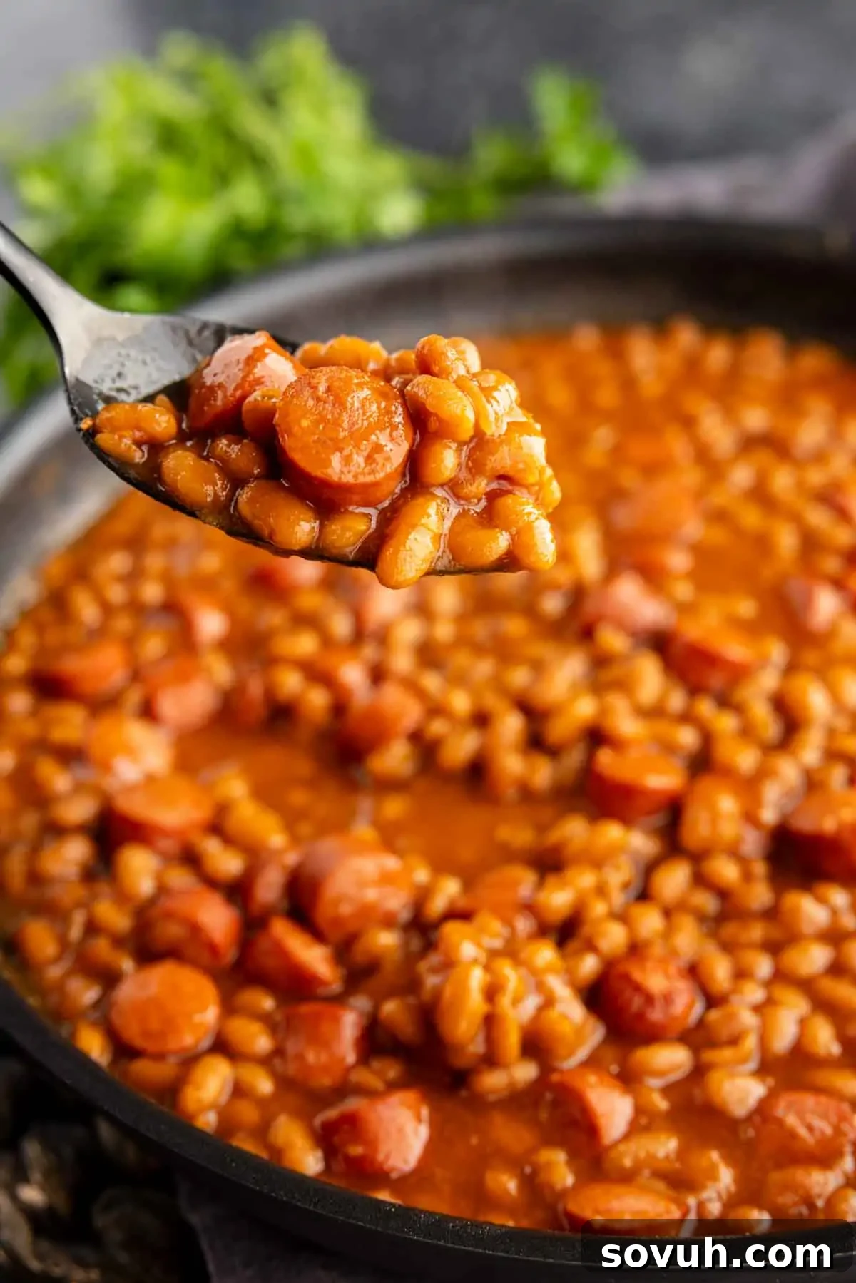 A spoon holds Homemade Franks and Beans over a pan filled with the same dish.  
