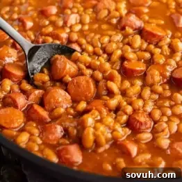 Close-up of baked beans mixed with sliced sausages in a pan, with a spoon scooping some.