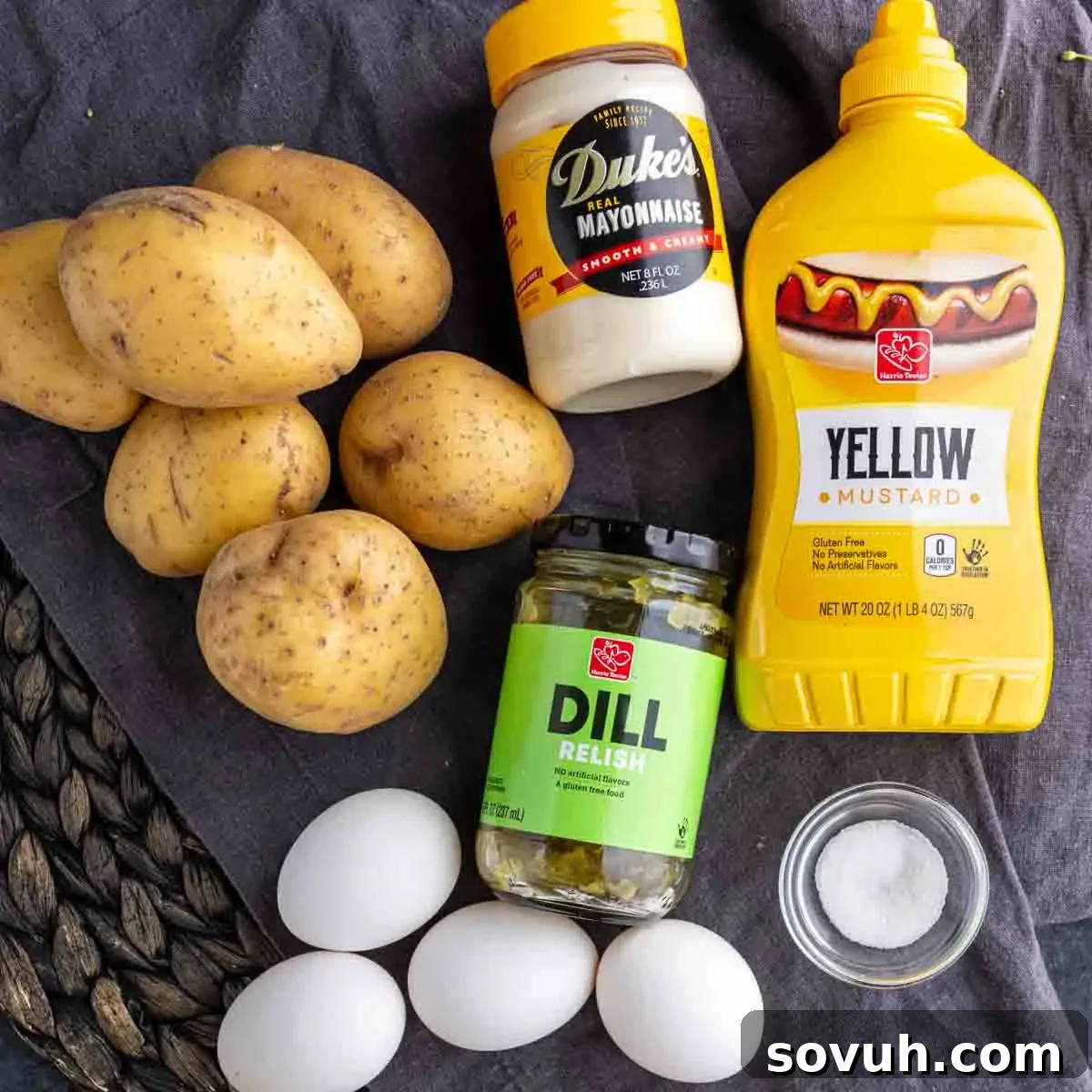 Potatoes, eggs, mayonnaise, yellow mustard, dill relish, and a small bowl of salt on a dark cloth.