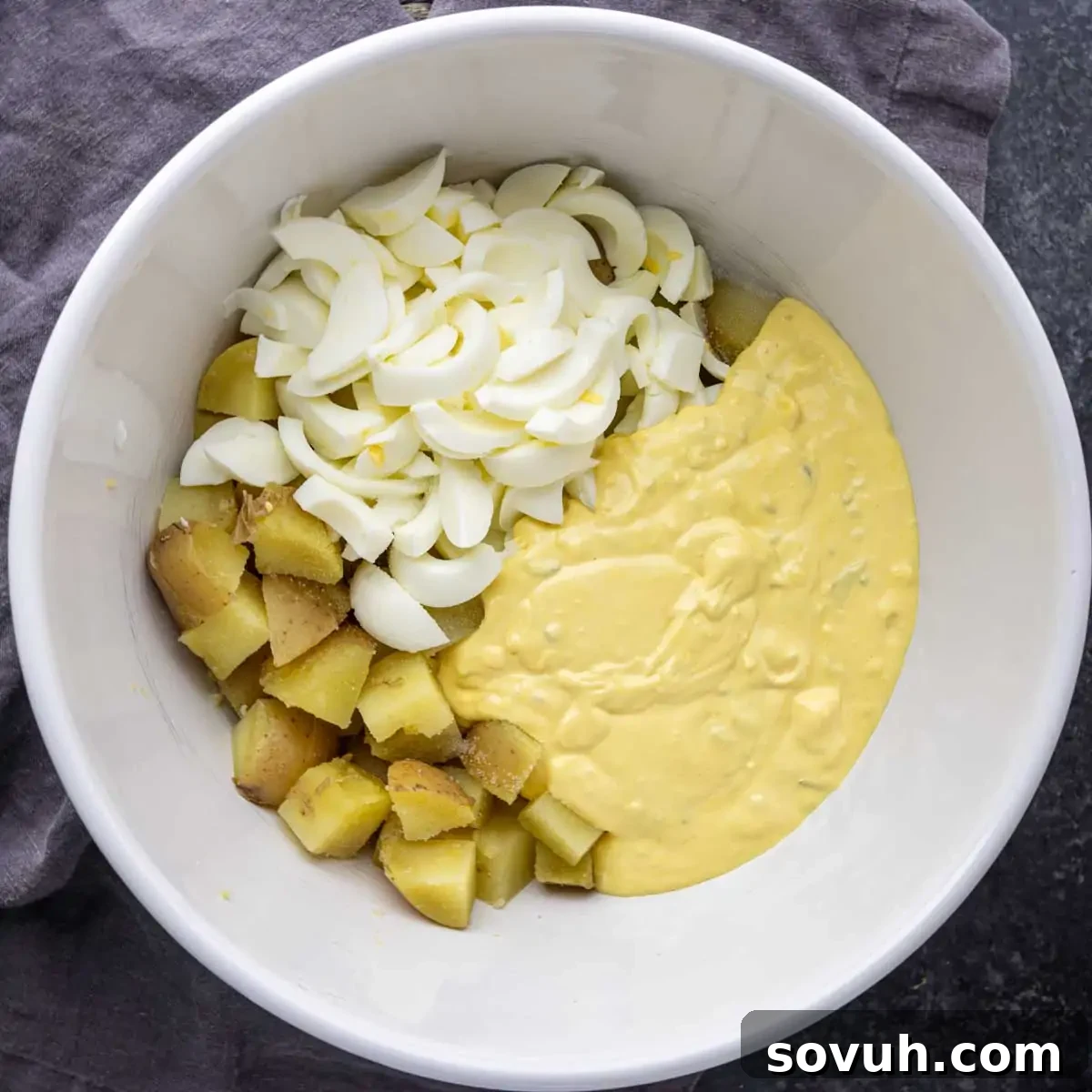 A white bowl containing chopped boiled eggs, diced potatoes, and yellow mayonnaise.