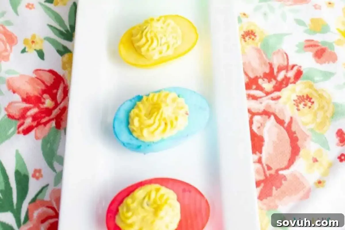 Three deviled eggs are colored in yellow, blue, and red with yellow filling, placed on a white rectangular plate over a floral tablecloth.