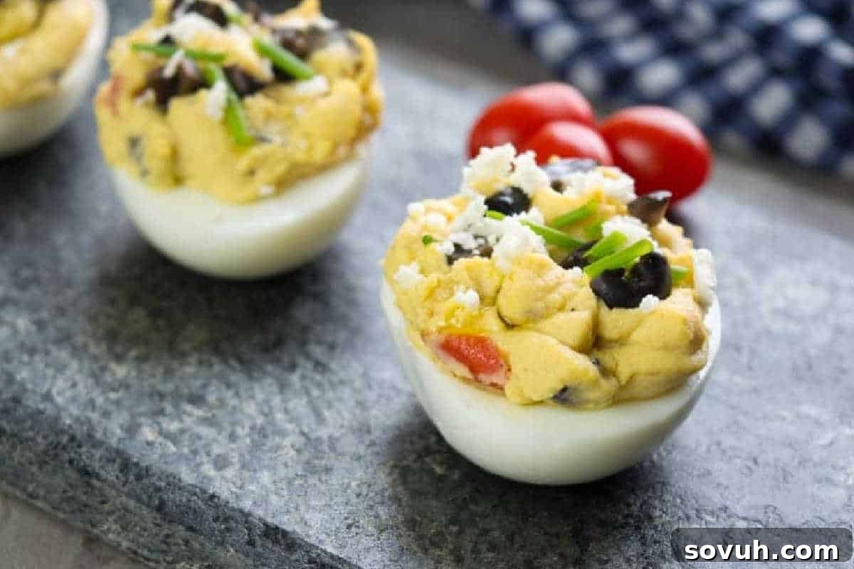 Stuffed egg halves with a creamy mixture, topped with olives, chives, and crumbled cheese. Cherry tomatoes are blurred in the background.