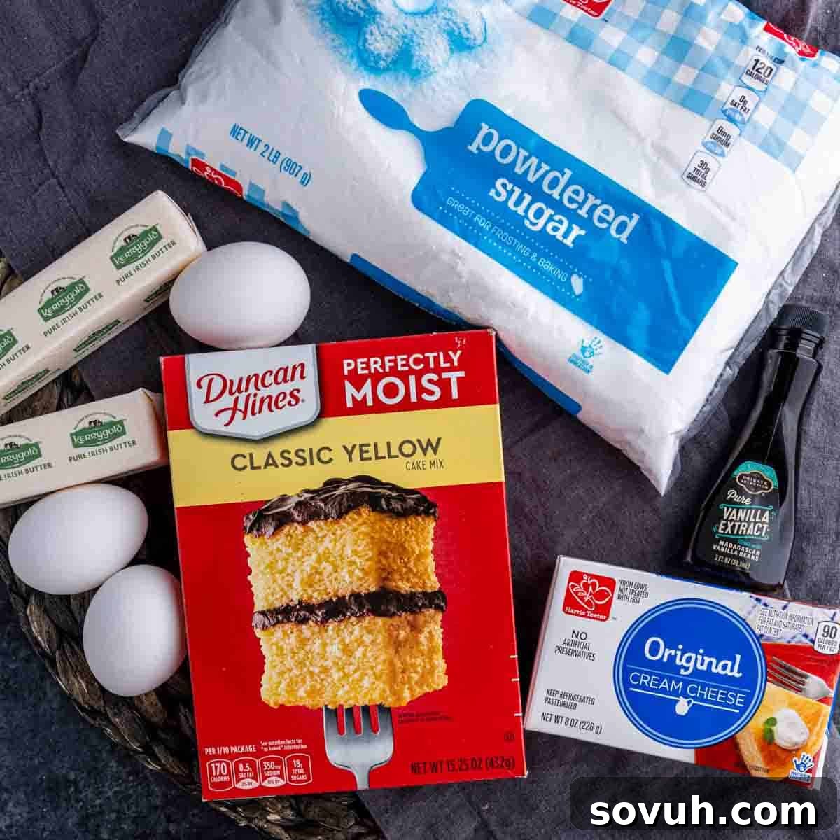 A collection of baking ingredients artfully arranged on a gray surface, including a box of yellow cake mix, a block of cream cheese, a bottle of vanilla extract, a pile of powdered sugar, several sticks of butter, and fresh brown eggs, ready for baking delicious Chess Squares.