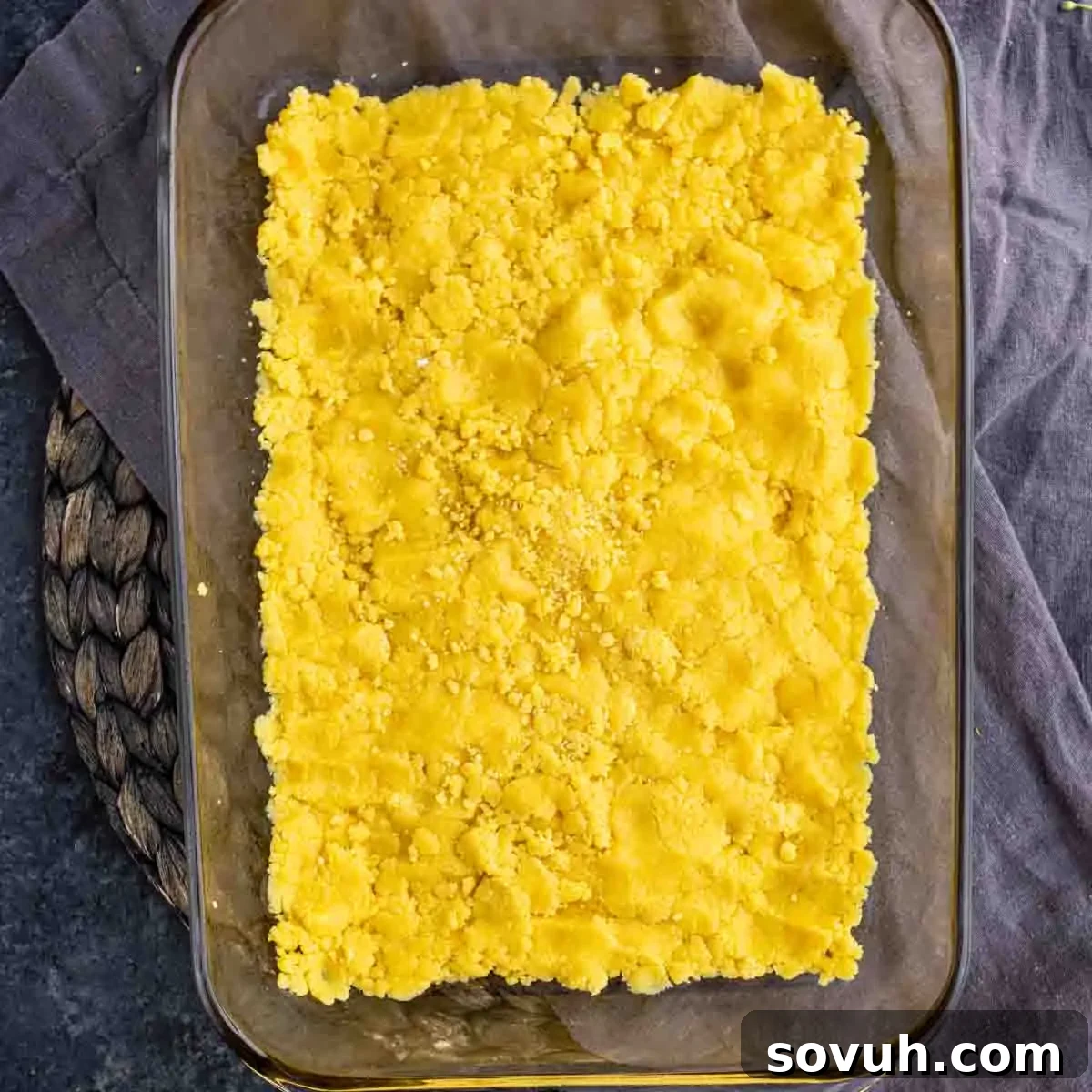 A rectangular baking dish contains a layer of raw yellow cake mix over a woven mat on a gray cloth, showing the initial step of preparing the base.