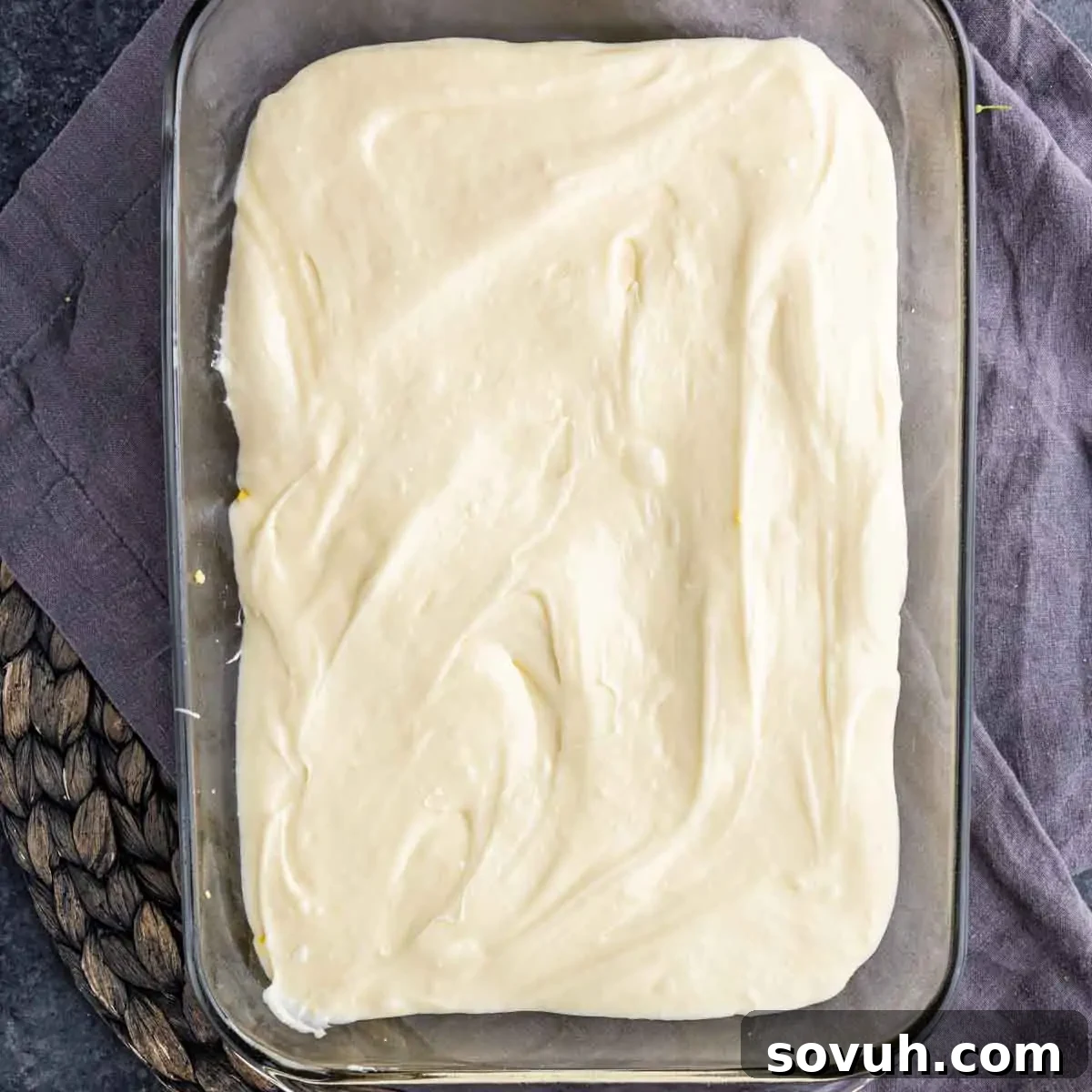 A rectangular glass dish filled with smooth, uncooked cake batter rests on a gray cloth, placed on a woven mat, illustrating the prepared base before the topping is added.