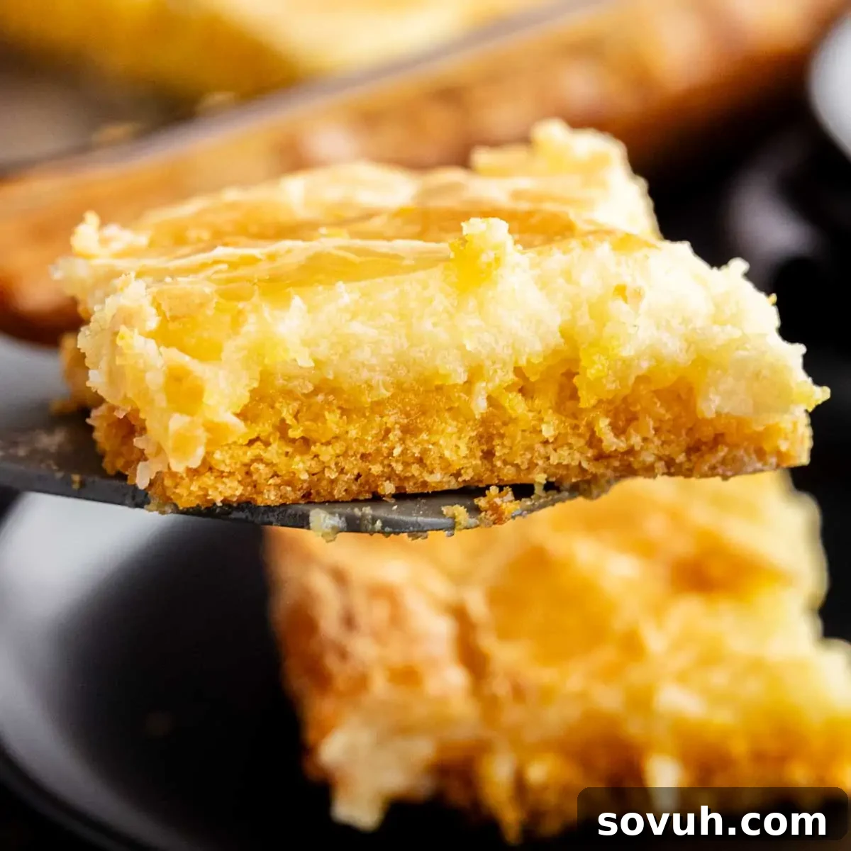 Close-up of a perfectly cut slice of layered Chess Square with a golden crumb base and creamy top, delicately held on a spatula above a black plate, showcasing its tempting gooey texture.