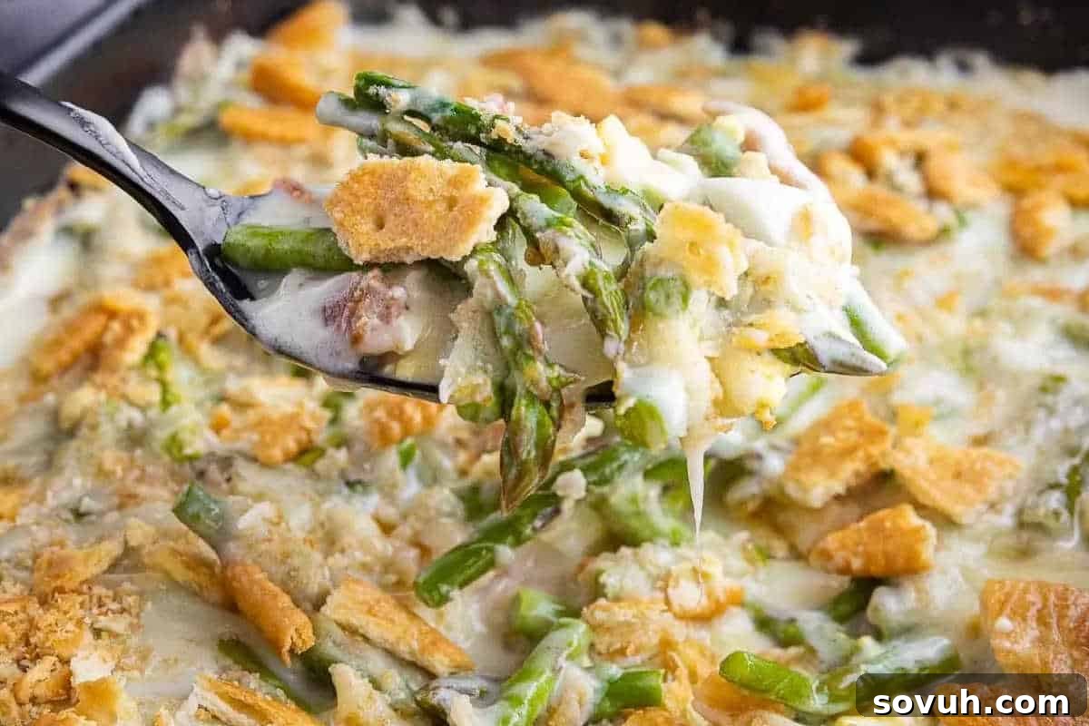 Close-up of a spoon lifting a portion of creamy asparagus casserole topped with crumbled crackers, cheese, and sauce in a baking dish.