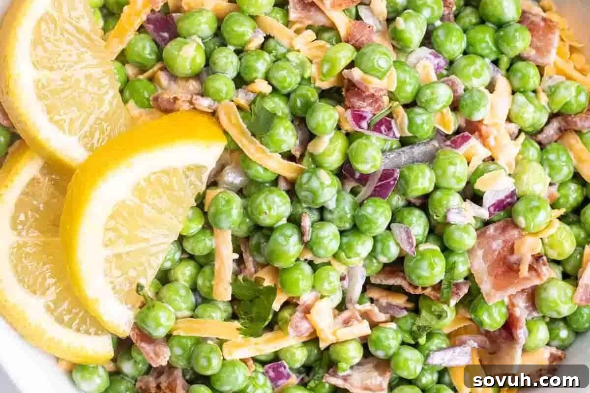 Close-up of a colorful pea salad with chopped red onions, crispy bacon, shredded cheese, and lemon slices on the side.