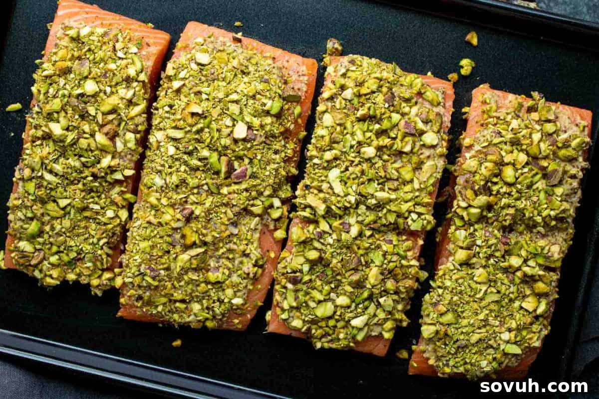 Four pieces of salmon are topped with a pistachio crust and arranged on a black baking tray.