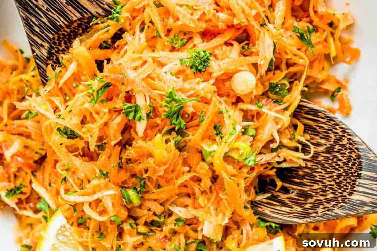 Close-up of a carrot salad with grated carrots, chopped parsley, and lemon slices, served in a wooden bowl with wooden serving spoons.