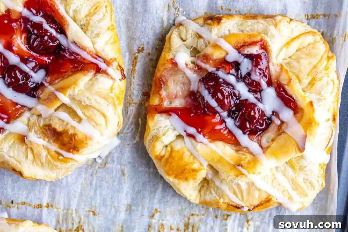 Two freshly baked cherry danishes with drizzled icing on a baking tray.