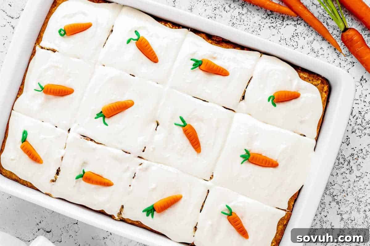A rectangular carrot cake with cream cheese frosting is cut into twelve squares, each topped with a small decorative carrot. Fresh carrots are visible in the top right corner.