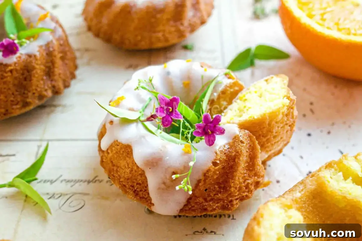 Mini bundt cakes with white icing and small purple flowers, garnished with green leaves, placed on a paper surface. An orange wedge is partially visible.
