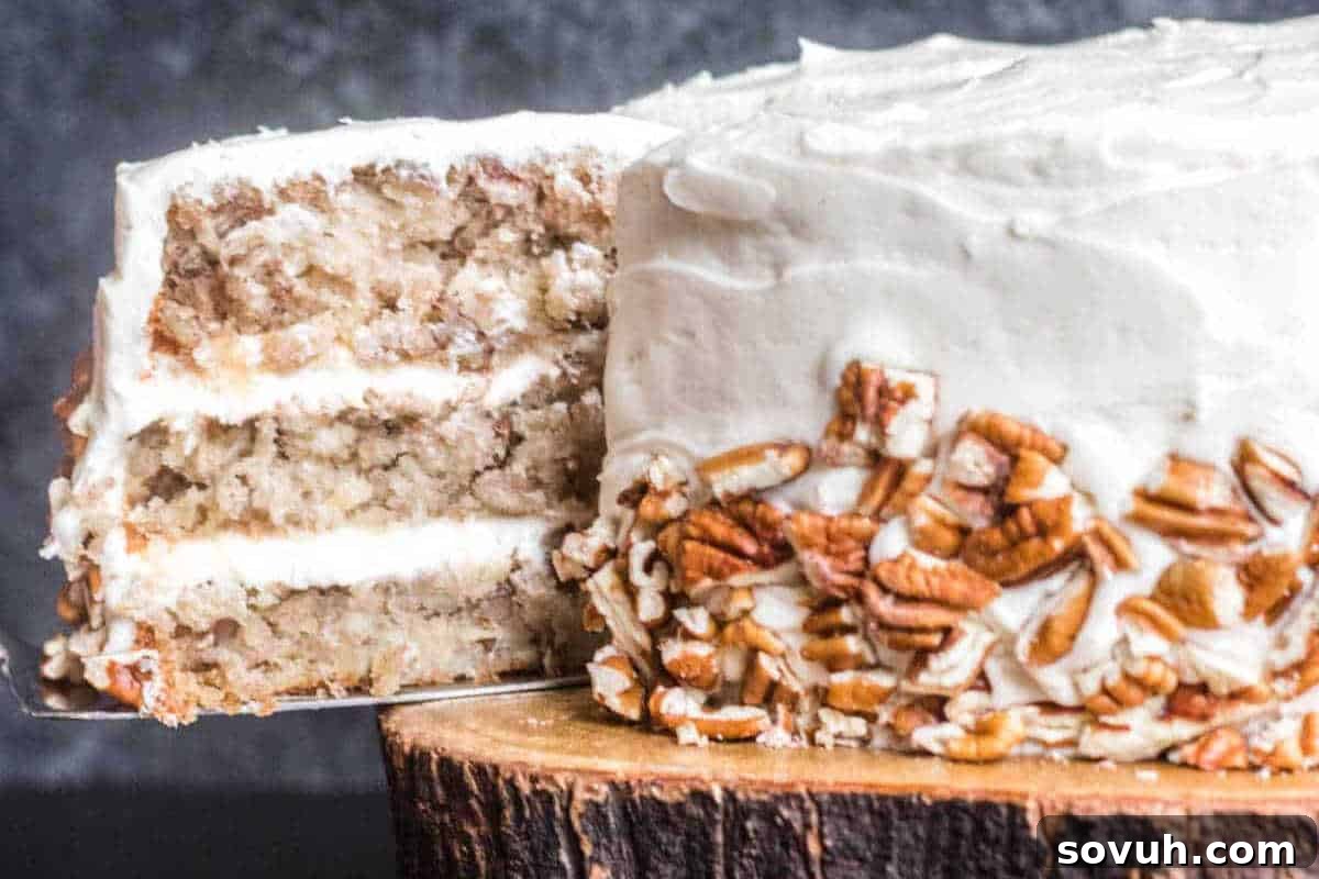 Three-layer cake with white frosting and pecans on the side, partially sliced, displayed on a wooden stand.
