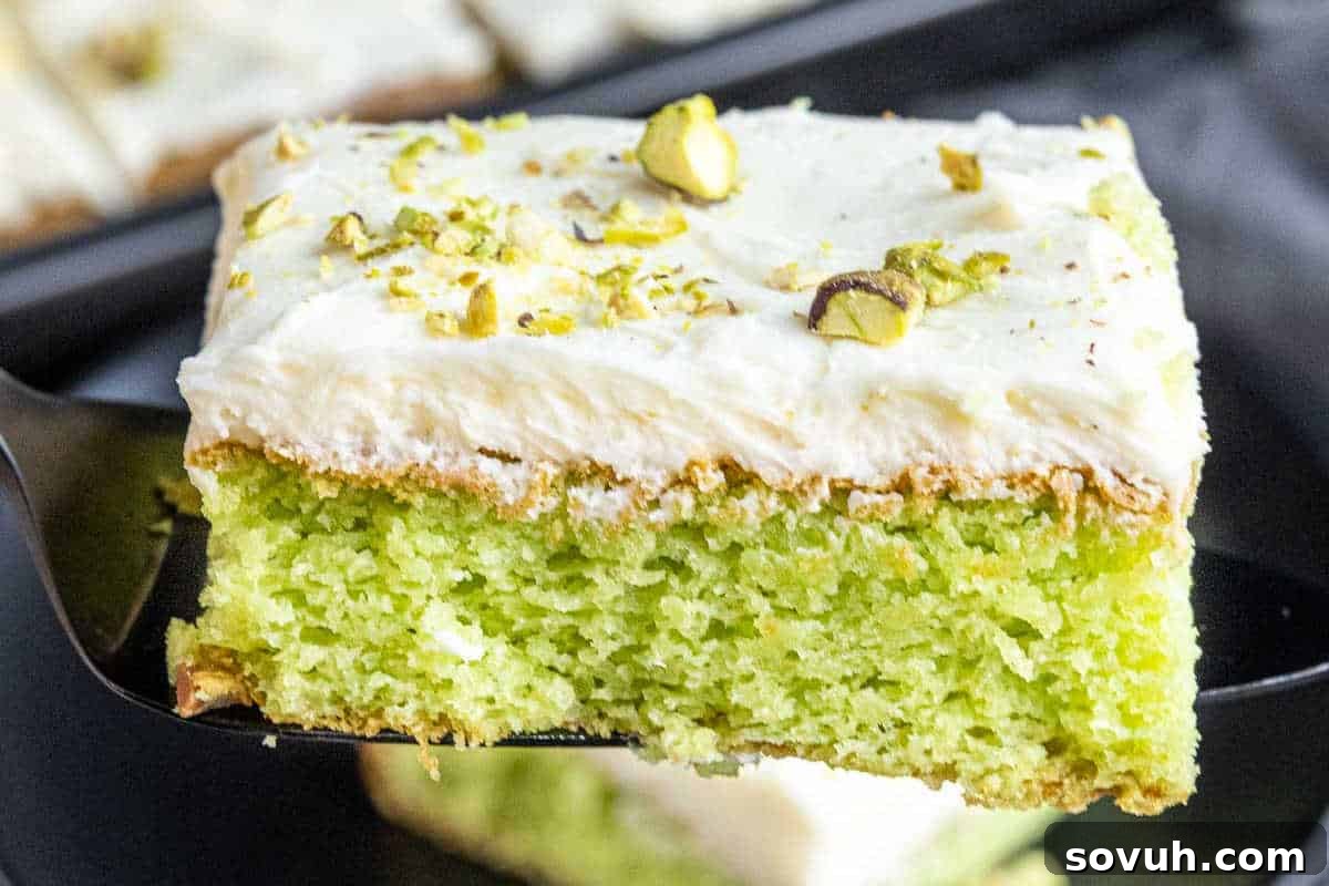 A slice of green cake with white frosting, garnished with chopped pistachios, being lifted on a spatula.
