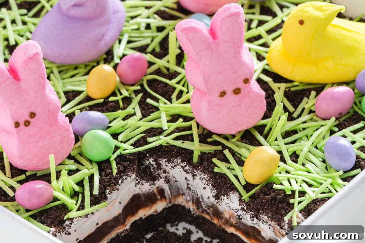Easter-themed dessert with pink, yellow, and purple marshmallow bunnies, green coconut grass, and colorful candy eggs on chocolate and cream layers in a dish.