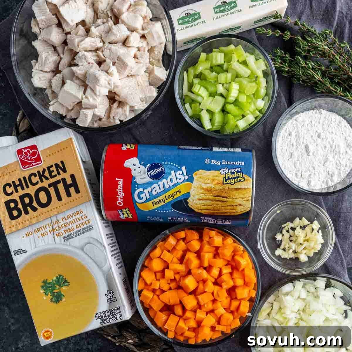 Ingredients for chicken pot pie, including diced chicken, celery, carrots, onion, garlic, flour, canned biscuits, chicken broth, butter, and thyme, arranged on a dark surface.