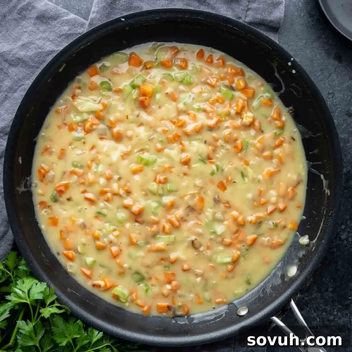 A skillet filled with a creamy mixture of diced carrots and celery, set on a dark surface with a gray cloth and parsley nearby.