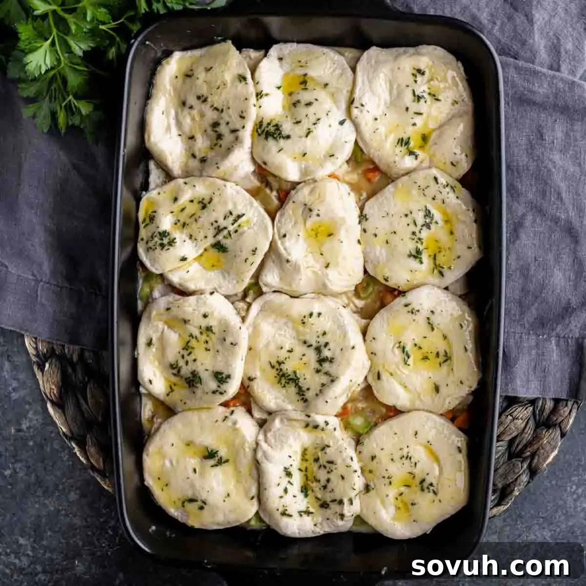 A baking dish with biscuits on top of a mixture containing vegetables and sauce, ready to be baked. Parsley garnishes the biscuits.