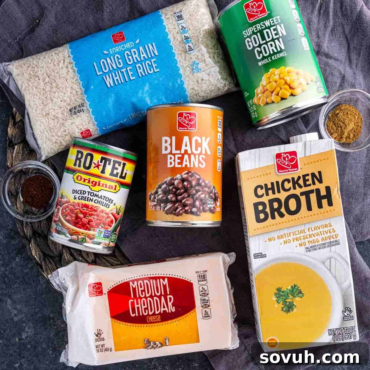 A selection of fresh and canned ingredients including long grain white rice, golden corn kernels, diced tomatoes with green chilies, black beans, chicken broth, medium cheddar cheese, and small bowls of spices.