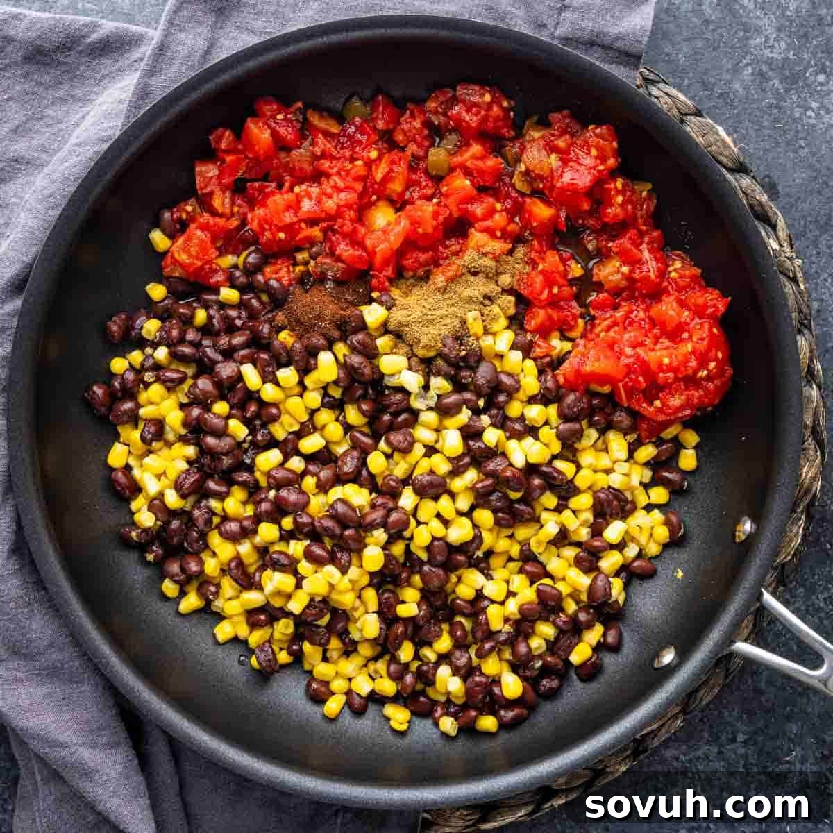 A black skillet containing a colorful mixture of black beans, corn, diced tomatoes with green chilies, and Southwest spices, placed on a dark countertop next to a gray dish towel.