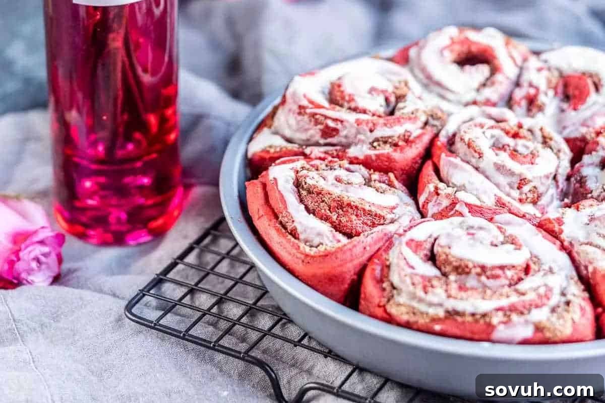 A round baking pan holds several freshly baked Red Velvet Cinnamon Rolls, each generously adorned with creamy white icing, resting on a cooling rack. To the left of the pan, a decorative bottle filled with a vibrant pink liquid adds a touch of charm to the scene.