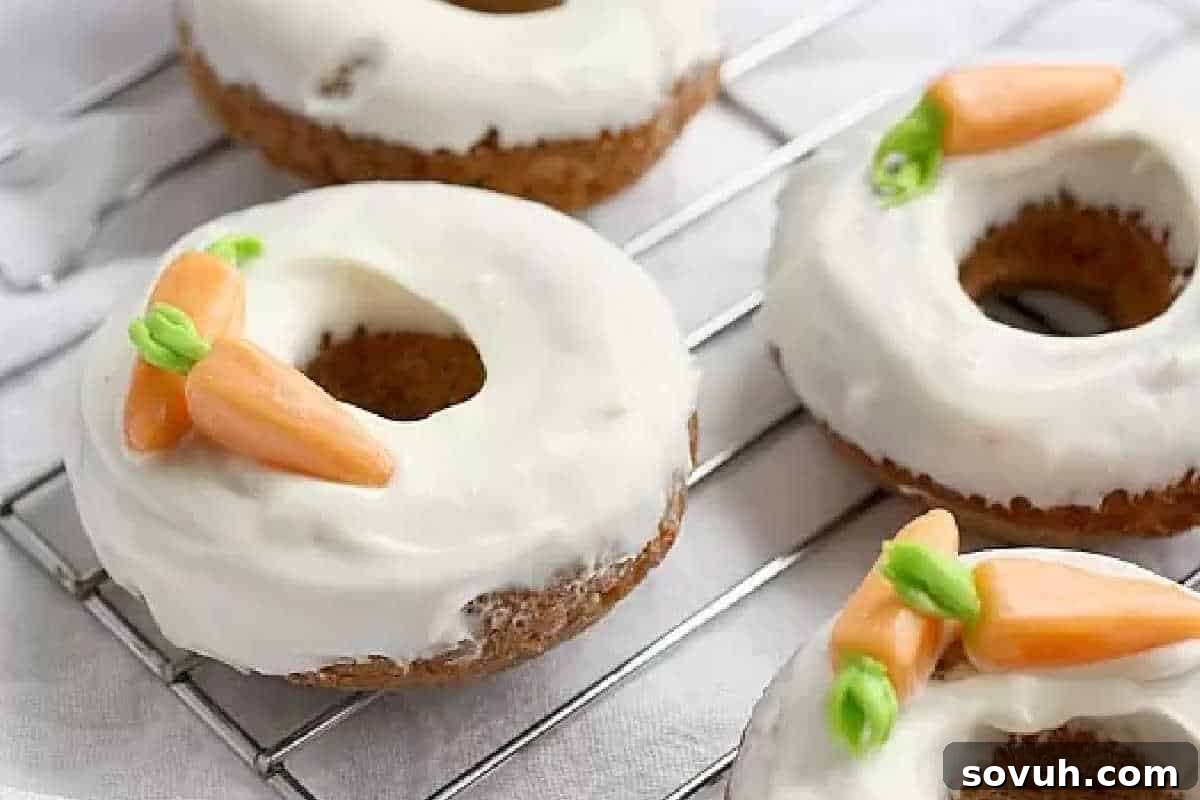 Four perfectly baked donuts, generously covered in smooth white icing, each charmingly topped with small, edible orange and green carrot-shaped decorations. These delightful treats are neatly arranged on a metallic cooling rack, presenting an appealing sight for fans of easy cake mix recipes eager to create impressive desserts with minimal effort.