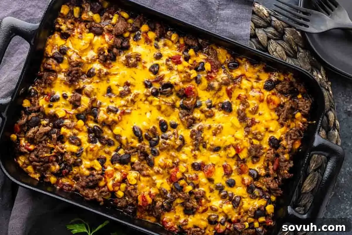 A baked casserole in a black dish, topped with melted cheddar cheese, beans, corn, ground beef, and diced tomatoes, on a woven placemat with plates and forks nearby.