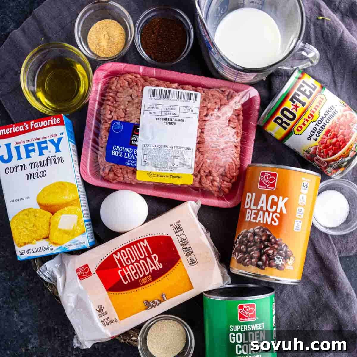 Assorted ingredients on a dark surface including ground beef, corn muffin mix, cheddar cheese, black beans, golden corn, Rotel tomatoes, an egg, milk, oil, and spices.