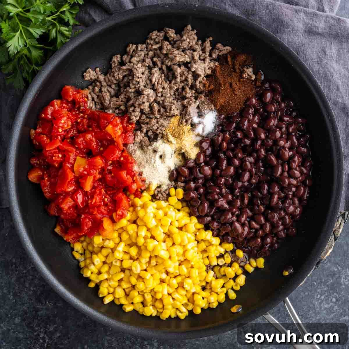 A skillet containing cooked ground beef, black beans, corn, diced tomatoes, and various spices, arranged in separate sections before mixing.