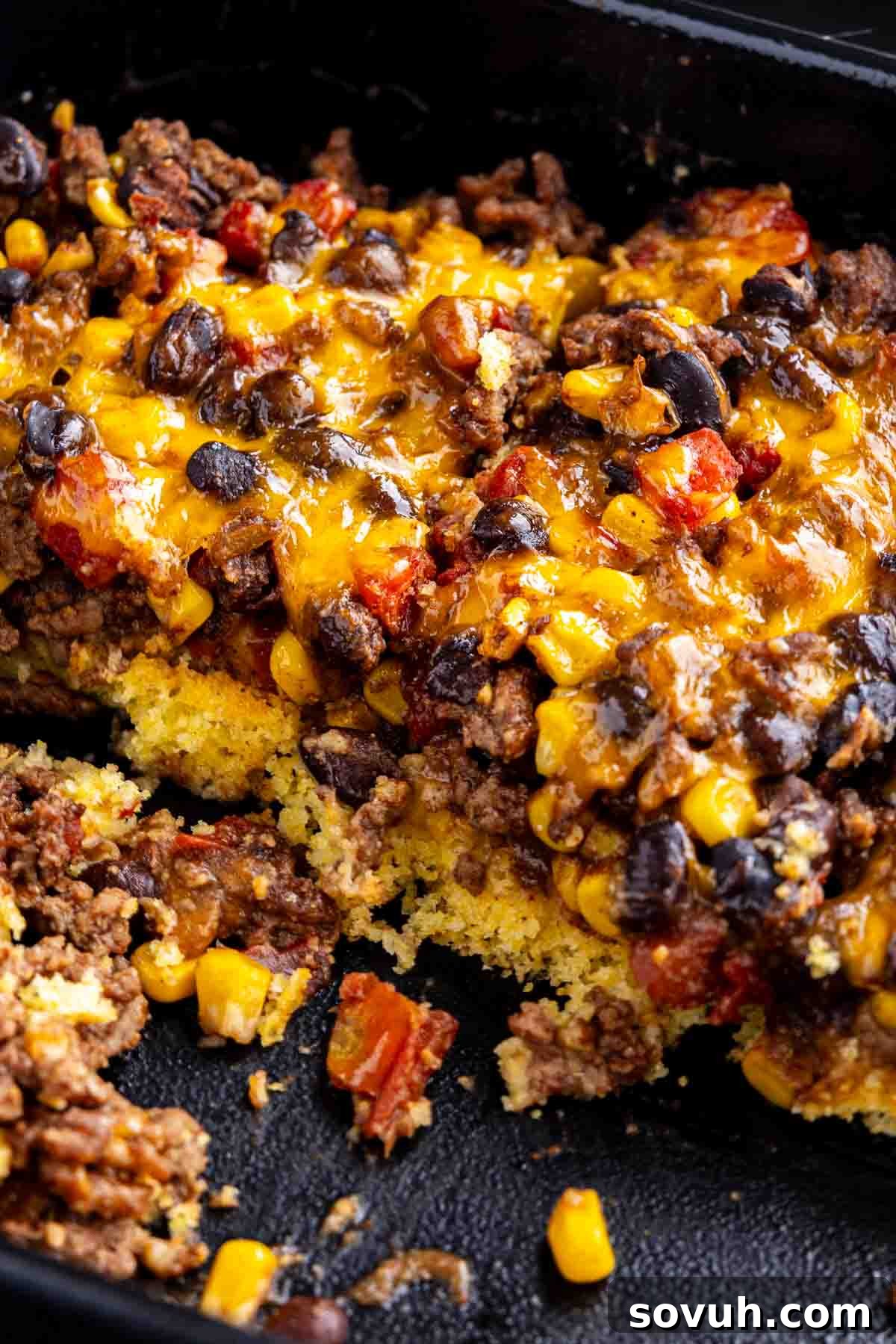 A close-up of a baked casserole with ground beef, black beans, corn, tomatoes, and melted cheddar cheese on a layer of cornbread in a black baking dish.