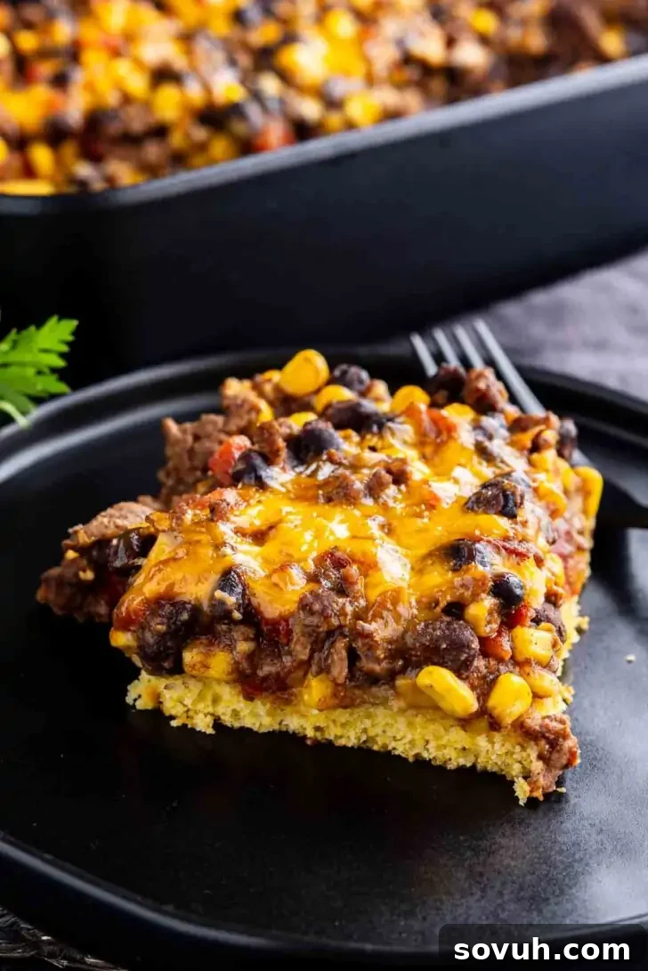 A slice of casserole with ground beef, black beans, corn, and melted cheese sits on a black plate, with the rest of the casserole in the background.