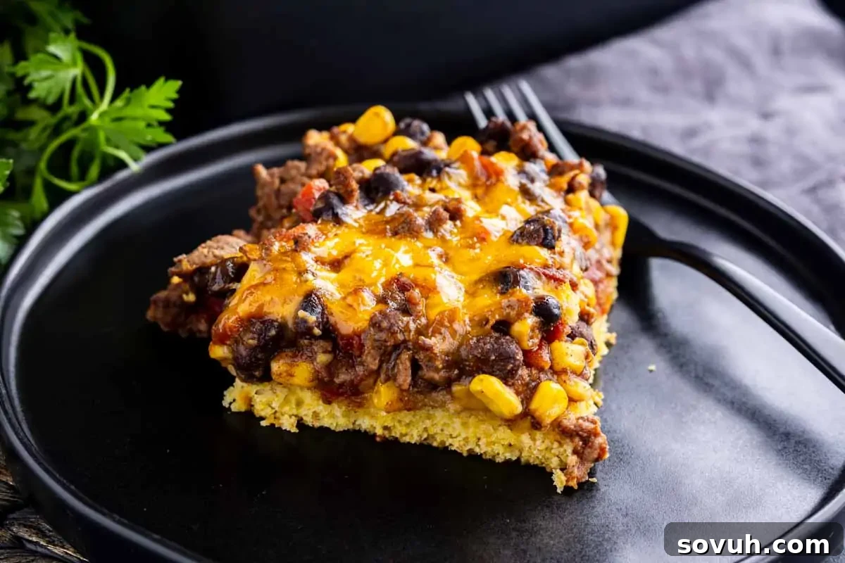 A generous serving of a hearty Black Bean Cornbread Casserole, richly topped with melted cheddar cheese, vibrant black beans, sweet corn, and savory ground beef. The dish sits invitingly on a sleek black plate, accompanied by a fork, showcasing its comforting appeal and tempting textures.