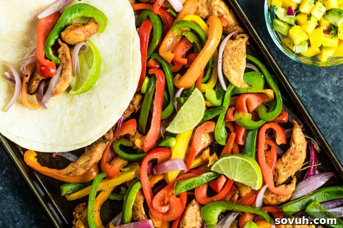 A vibrant and appetizing Sheet Pan Chicken Fajitas dish, featuring perfectly cooked, seasoned chicken alongside colorful bell peppers, tender onions, and bright lime slices, all arranged on a single baking sheet. The meal is complemented by a soft tortilla and a side of fresh mango salsa, promising a complete and flavorful experience.