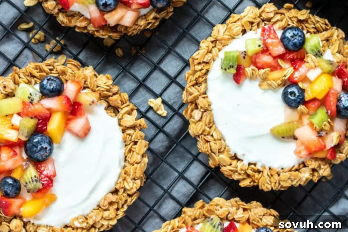 Three granola cups filled with yogurt and topped with assorted fresh fruits, on a dark grid background.