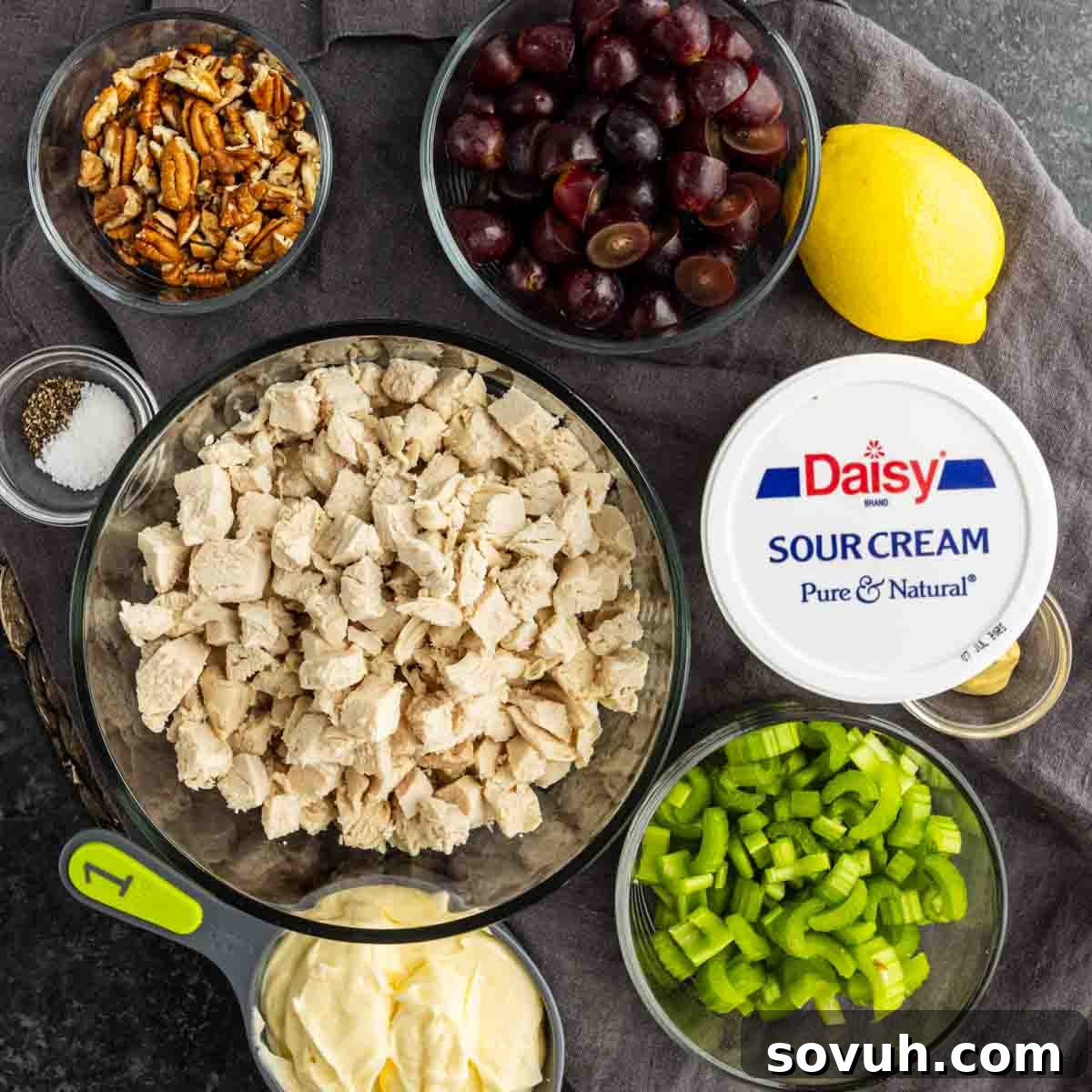 Sweet and Savory Chicken Grape Salad 3 Bowls with diced chicken, sliced grapes, chopped celery, pecans, mayonnaise, sour cream, a lemon, salt, and pepper arranged on a dark surface.