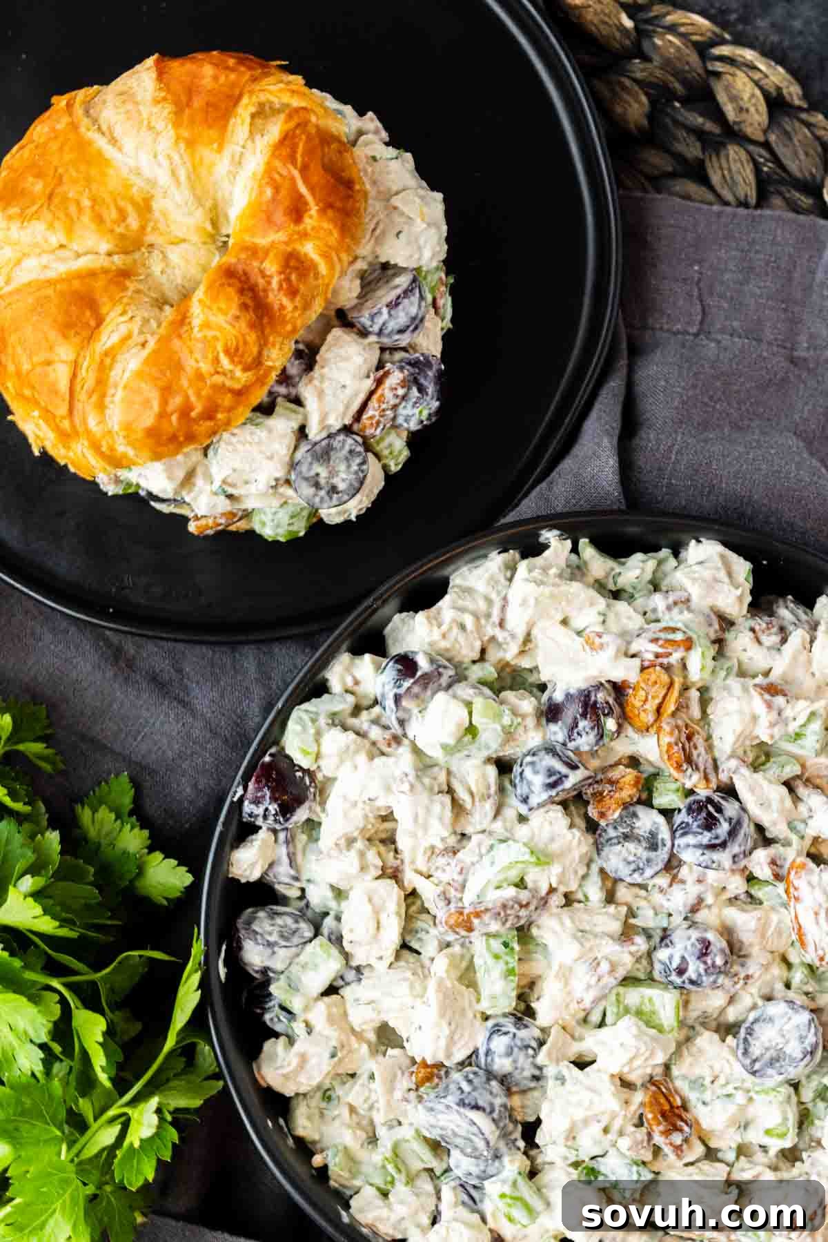 Sweet and Savory Chicken Grape Salad 6 A black plate with a croissant filled with chicken salad next to a bowl of chicken salad with grapes, celery, and pecans in a creamy dressing. Parsley is also visible.