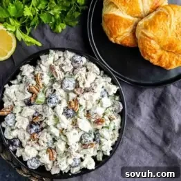 Sweet and Savory Chicken Grape Salad 7 A bowl of chicken salad with grapes, pecans, and celery sits next to a plate with two croissants on a dark tablecloth. Fresh parsley and a lemon wedge are also visible.