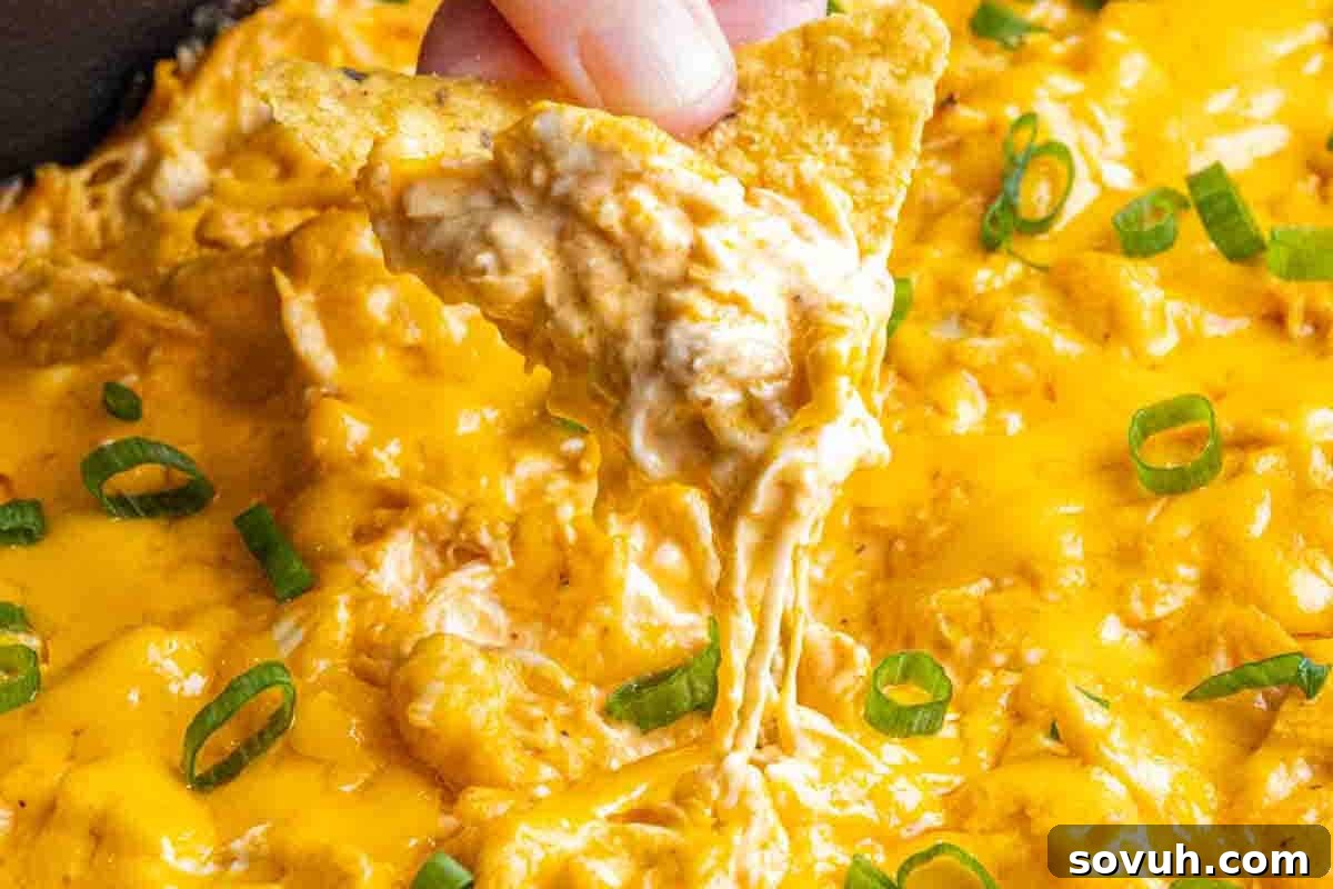 A hand dips a crisp tortilla chip into a bubbling, cheesy buffalo chicken dip, generously topped with melted cheese and vibrant sliced green onions, promising a perfect scoop every time.